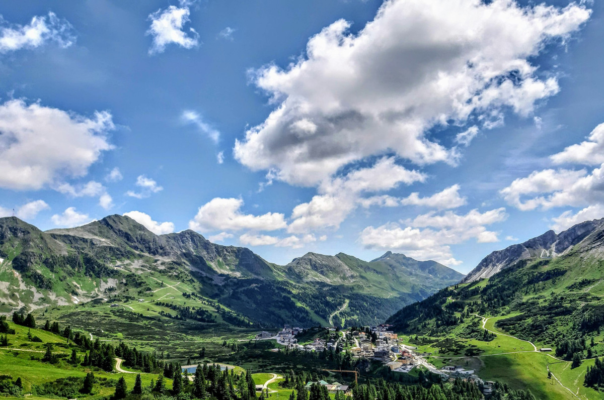 Panoramablick auf ein Bergdorf inmitten grüner Almwiesen und imposanter Berggipfel unter blauem Himmel mit weißen Wolken.