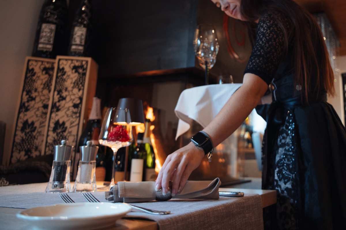 Eine Service-Mitarbeiterin in eleganter Kleidung deckt aufmerksam einen Restauranttisch ein. Im Vordergrund liegen Besteck, Teller und ein gefaltetes Stoffserviette. Das Bild vermittelt Professionalität und Liebe zum Detail im Service