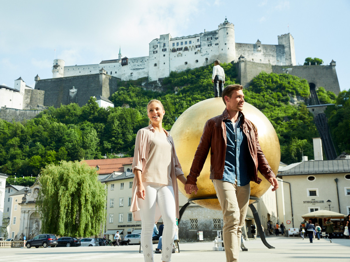 © SalzburgerLand Tourismus Paar spaziert vor der Festung Hohensalzburg und der goldenen Kugel am Kapitelplatz in Salzburg.
