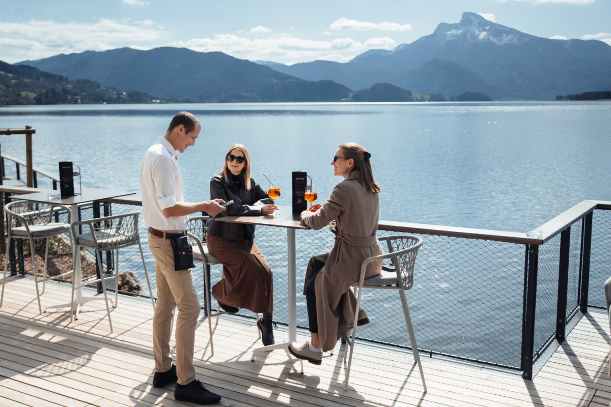 Two women enjoy Aperol drinks on a sunny lakeside terrace in the Salzkammergut as a waiter takes their order; in the background, a lake shimmers against a scenic mountain backdrop.