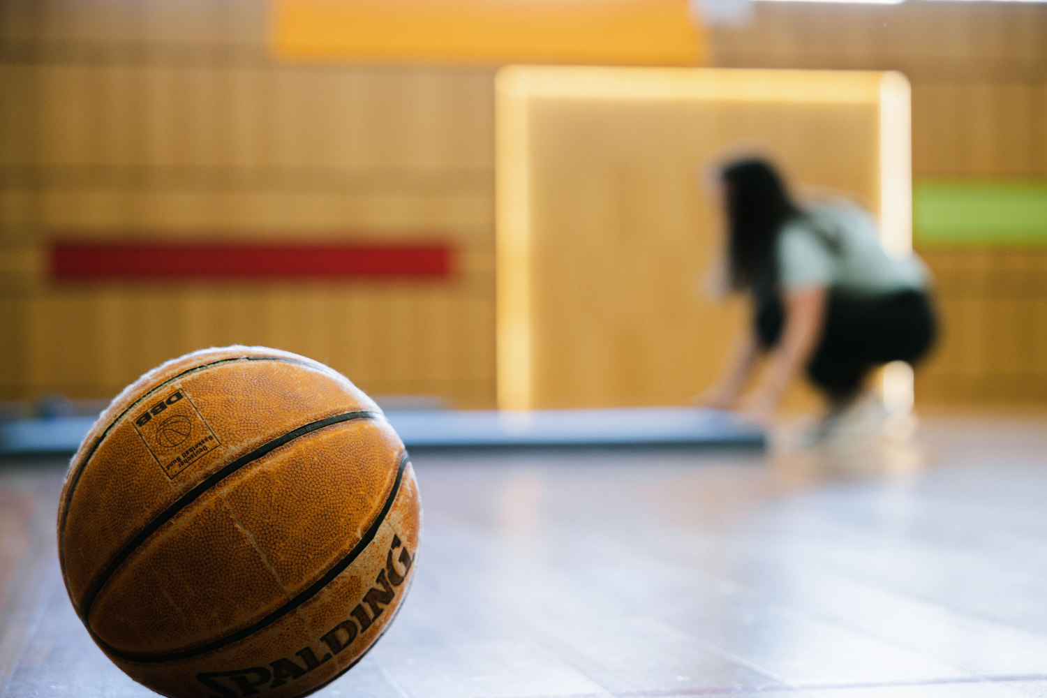 Ein Basketball im Vordergrund, eine Kinderbetreuuering im Hintergruns legte eine Matte in einem Turnsaal auf den Boden.