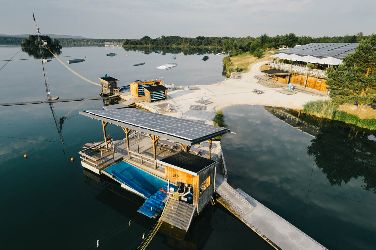 Luftaufnahme des Wild Wake Park in Bayern mit Wakeboard-Anlage auf einem ruhigen See, mehreren Hindernissen im Wasser