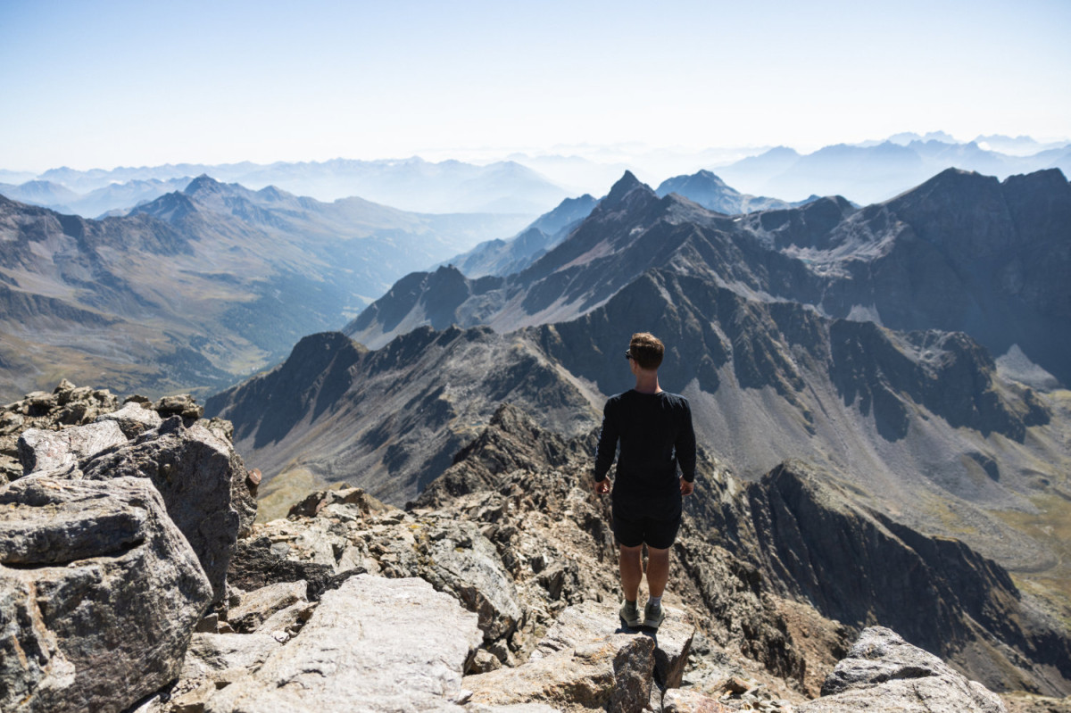 © Satellite Creative House Person steht auf einem felsigen Berggipfel und blickt über eine weite, zerklüftete Gebirgslandschaft mit mehreren Bergketten im Dunst.