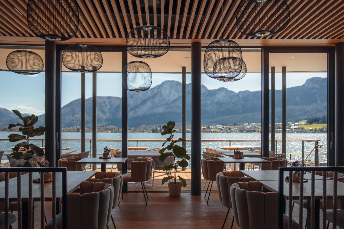 Interior view of a stylish restaurant overlooking lake in the Salzkammergut; modern decor with hanging lights and cozy chairs, with the shimmering water and alpine mountains in the background.