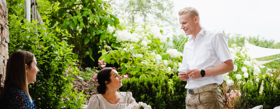 Junger Kellner bedient zwei Damen in einem Gastgarten.