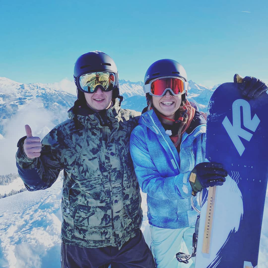 Zwei Snowboarder stehen auf der Piste und lachen in die Kamera