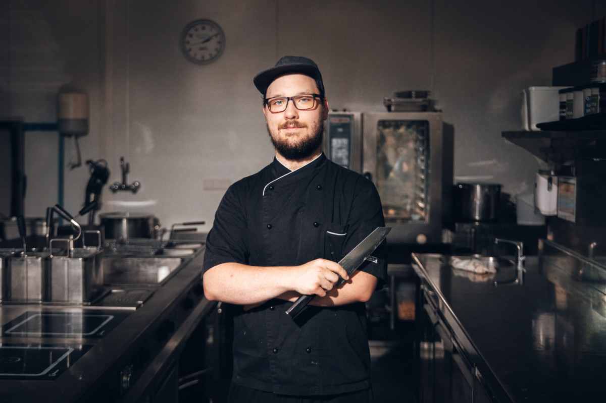 Koch mit schwarzer Schürze und einem Messer in der Hand steht in einer Industrieküche.