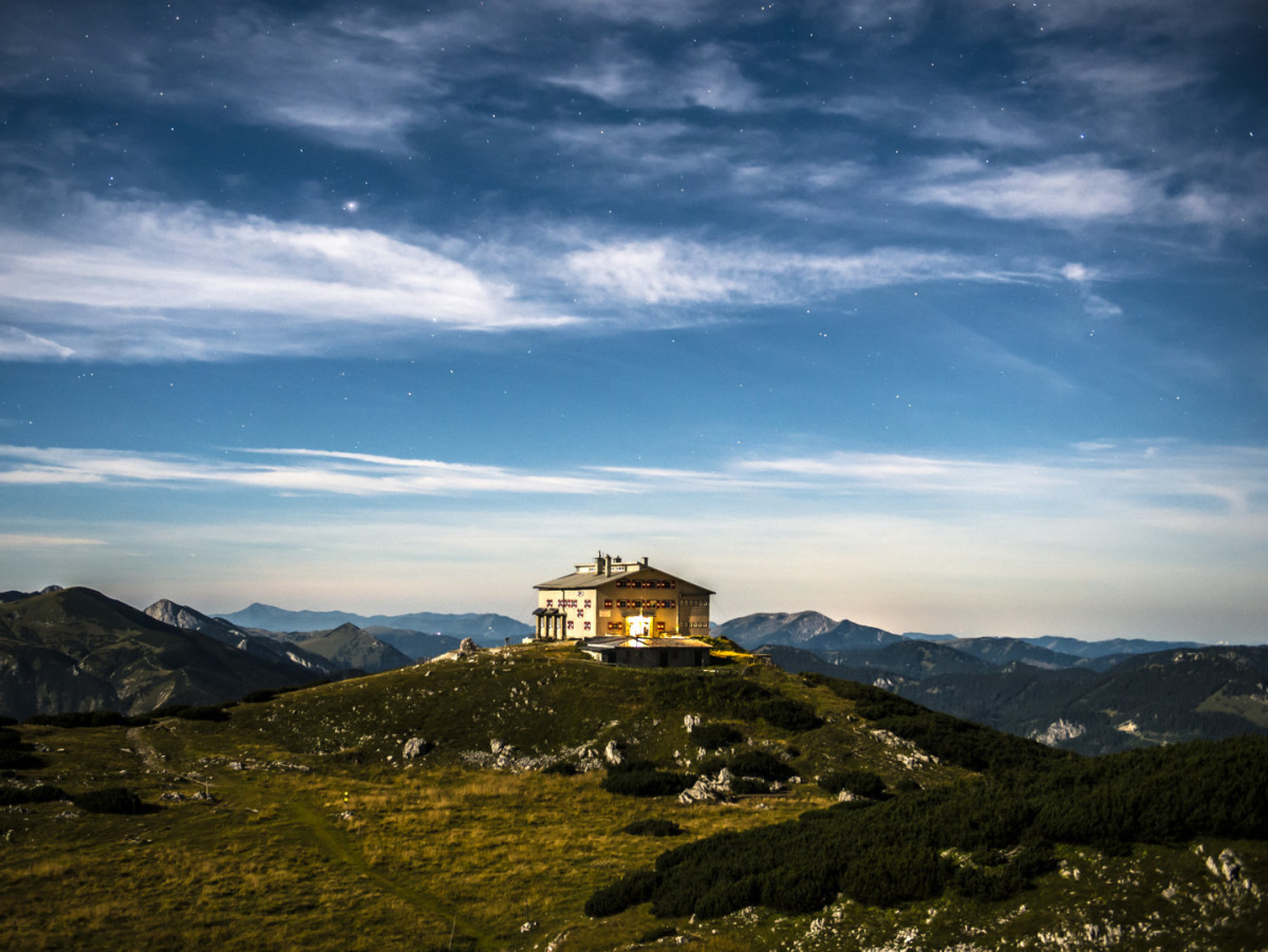 Berghütte auf einem Gipfel bei Nacht mit Blick auf die umliegende Berglandschaft und den Sternenhimmel.