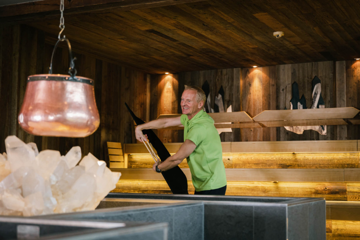 Ein Hotelmitarbeiter mit einem grünen T-Shirt bereitet in einer stilvollen Sauna mit Holzwänden einen Aufguss vor.