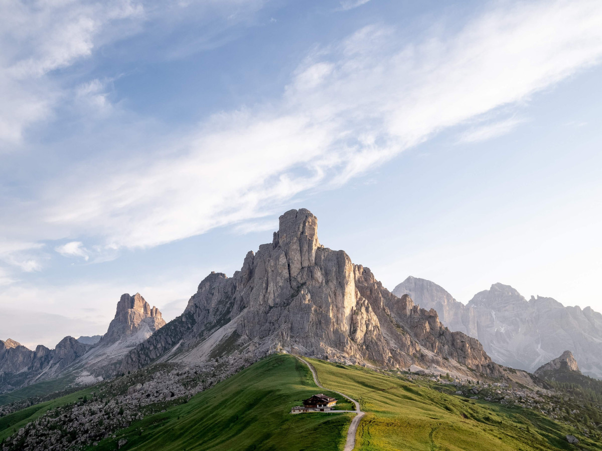 © Unsplash, Nicola Pavan Markante Felsgipfel der Dolomiten mit grüner Almwiese, Berghütte und Wanderweg.