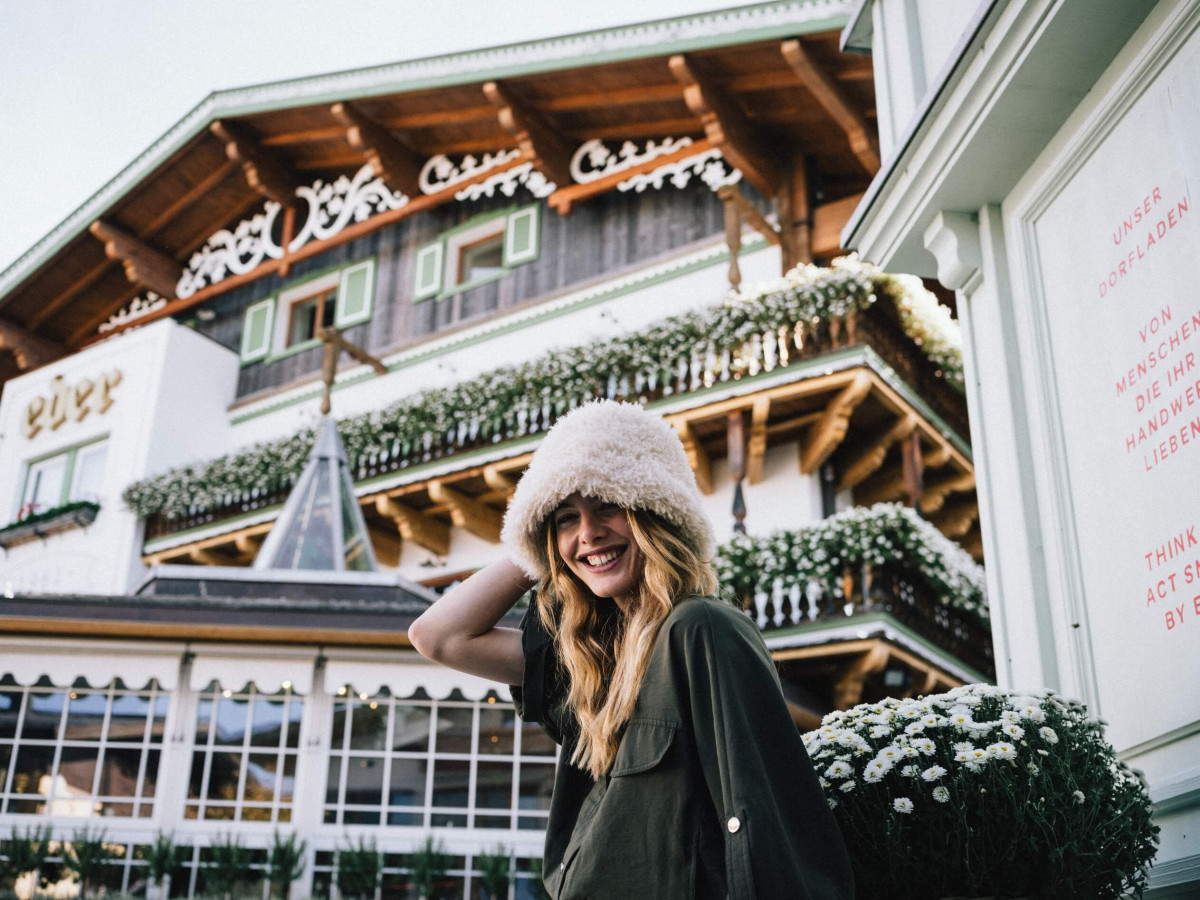Lachende Frau mit Wintermütze steht vor einem traditionellen Hotel der Eder Collection mit Holzbalkonen und Blumenschmuck.