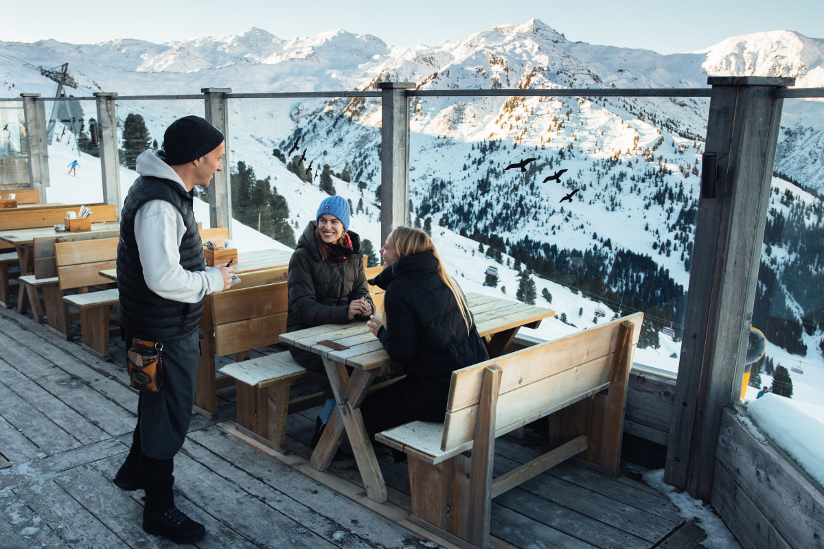 Ein Kellner bedient zwei Frauen auf der Terrasse einer Skihütte.