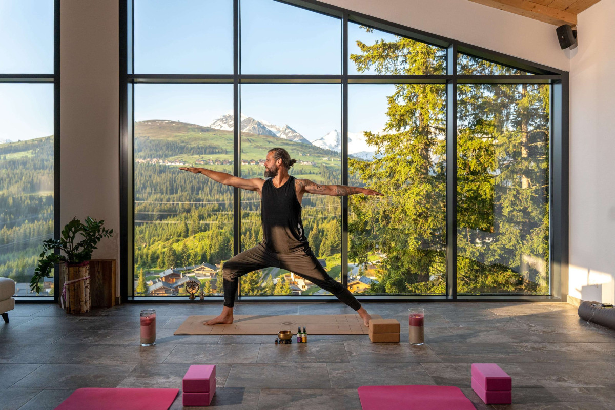 Mann macht die Yoga vor eine Glasfront im Hotel Ursprung, die den Blick auf eine atemberaubenden Bergkulisse freigibt. 