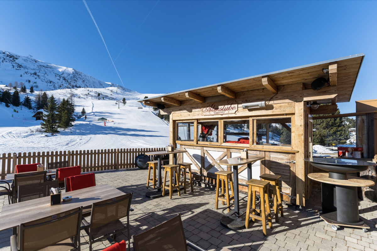 Terrasse des Gasthofs Almstube mit Holzhütte und Sitzplätzen, umgeben von schneebedeckten Hängen und blauem Himmel.
